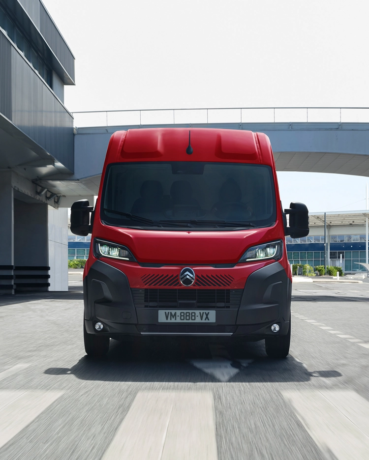 Vista frontal de una Citroën Jumper roja circulando por un entorno urbano moderno, destacando su diseño robusto, practicidad y fuerte presencia en carretera.