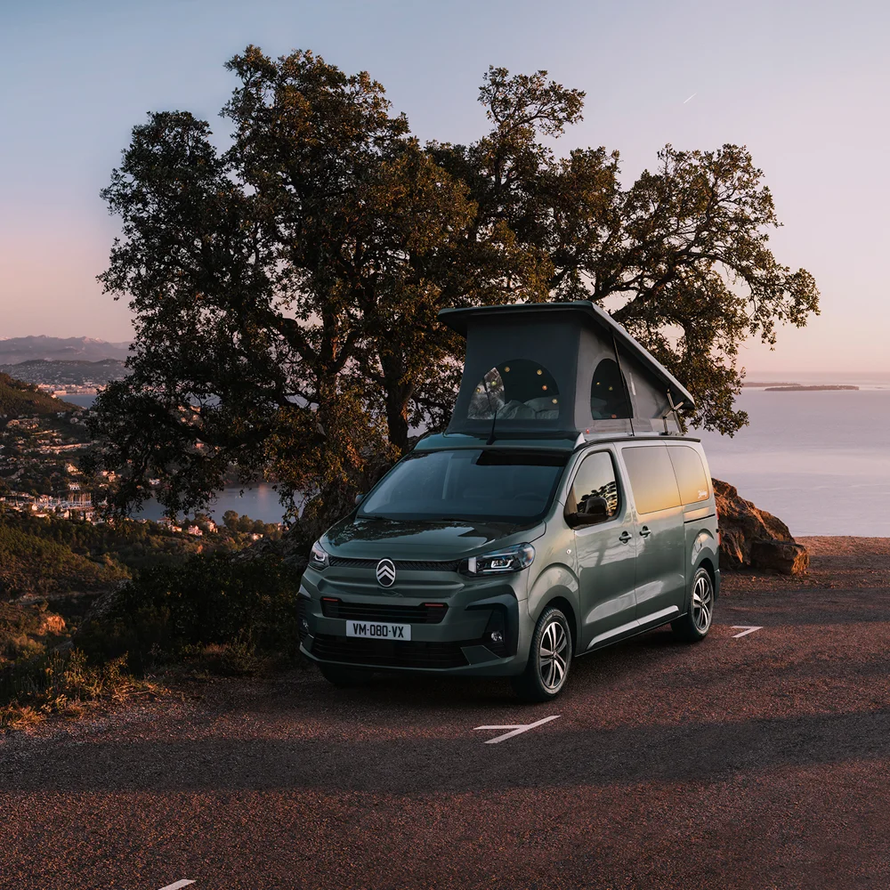 Furgoneta camper Citroën aparcada en una carretera costera con vistas al atardecer, con árboles y colinas al fondo