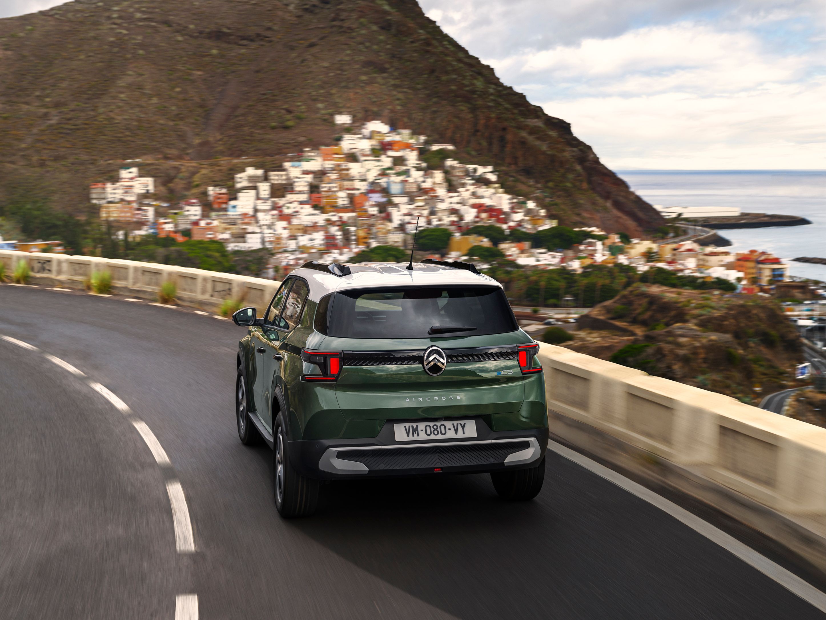 Vista trasera tres cuartos en conducción del Citroën ë-C3 Aircross SUV en una carretera costera para una movilidad diaria con confianza, suave y asequible.